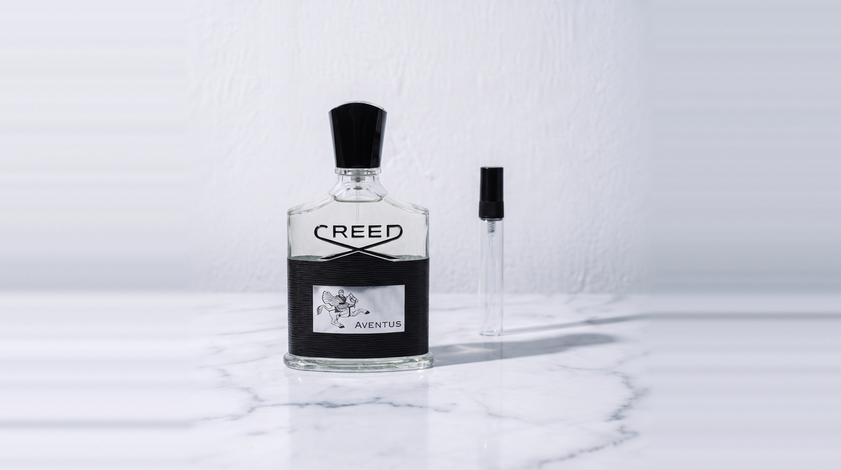 Botella original de Creed Aventus junto a un decant de vidrio sobre mármol blanco