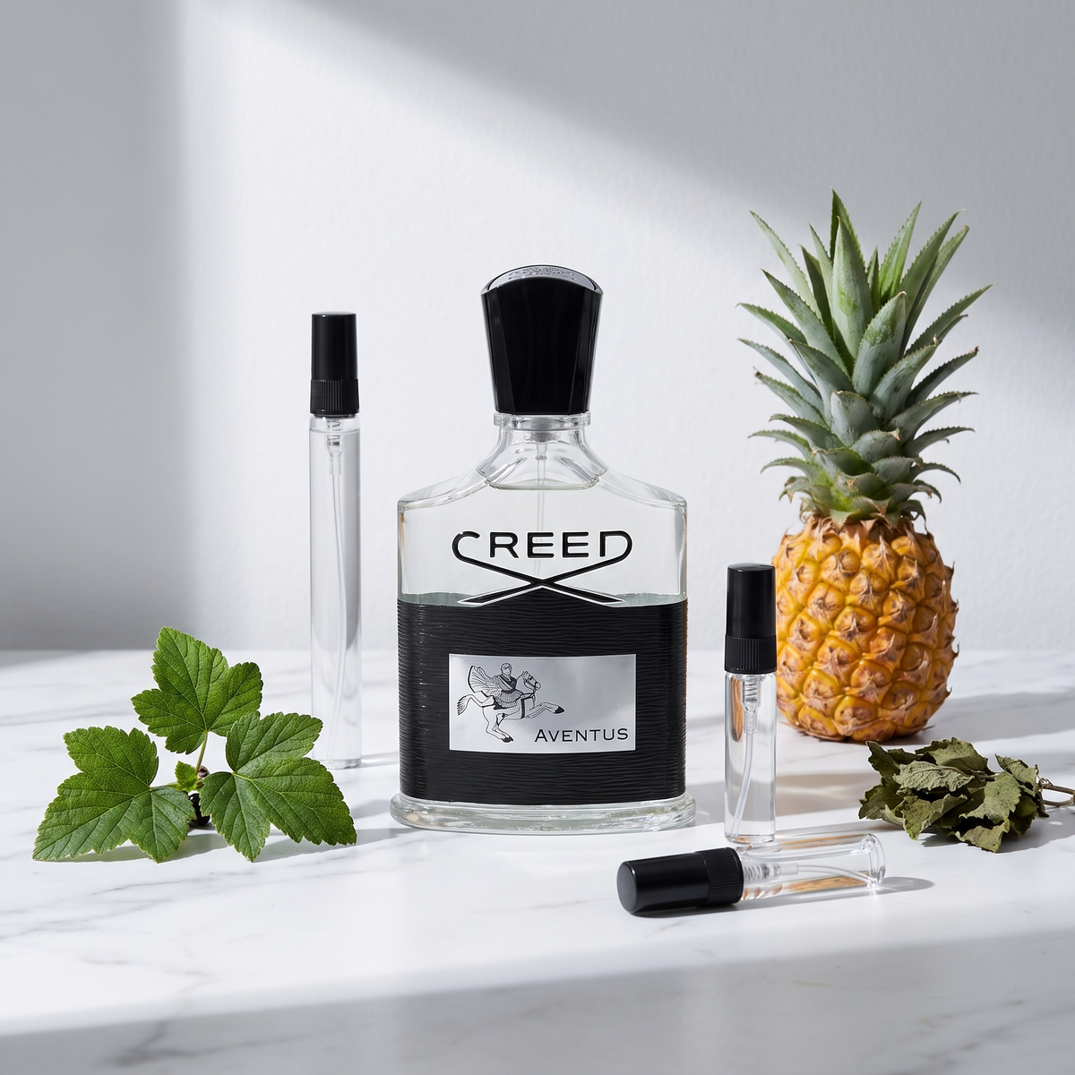 Decant de Creed Aventus en 3, 5 y 10 ml junto a la botella transparente original de Creed con una piña entera, hojas de grosella negra y hojas secas de pachulí